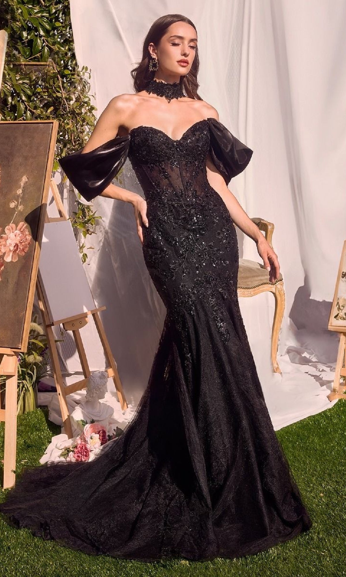 A woman poses outdoors among art easels and draped white fabric, wearing the Andrea & Leo A1354 long mermaid prom dress featuring a beaded evening gown with a sheer bodice and dramatic detached sleeves.